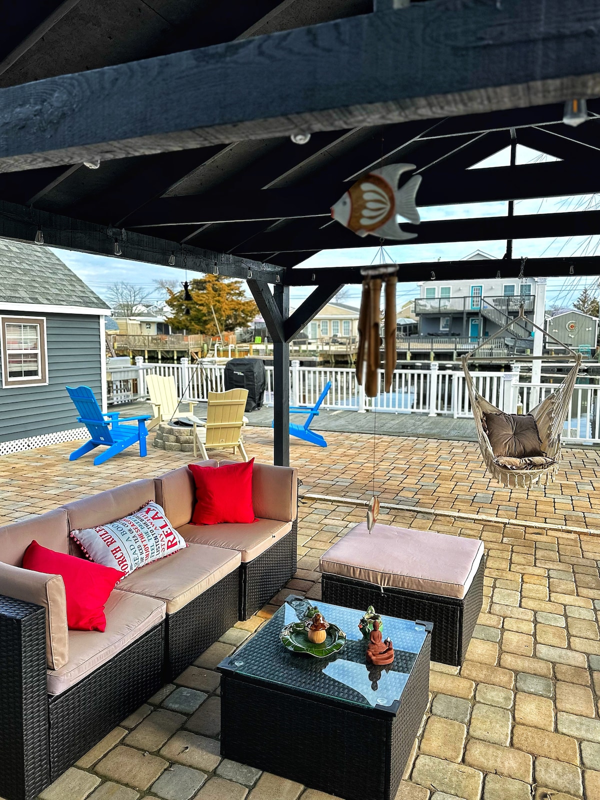 A covered gazebo is presented with a comfortable seating area featuring a dark wicker sofa adorned with red cushions. A glass-top coffee table sits in front, surrounded by colorful adirondack chairs on the patio paved with stones.