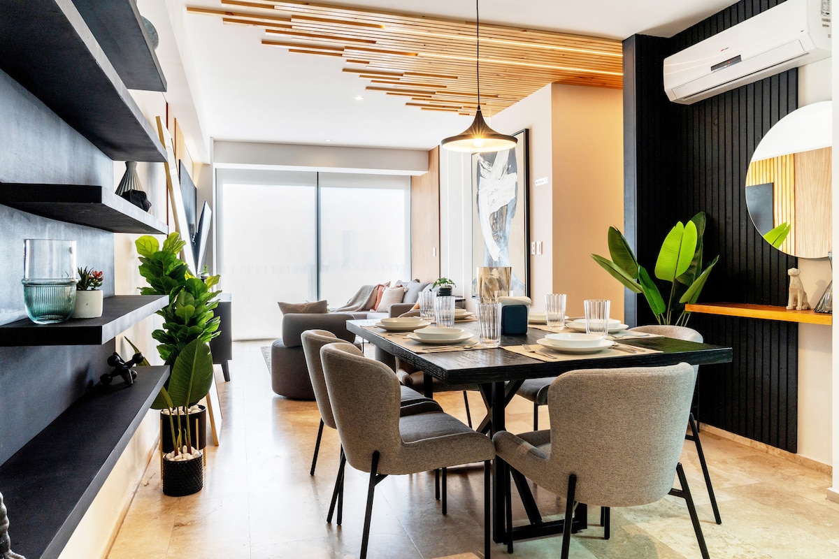 A modern dining area is presented, featuring a large black table surrounded by several grey upholstered chairs. Natural light is filtered through a large window, illuminating decorative greenery placed nearby. Unique ceiling beams add a touch of elegance, while an accent mirror reflects the room's design.