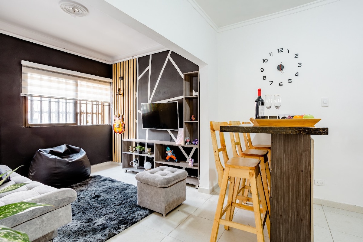 A modern living space is presented with a dark accent wall and a soft area rug. A television is mounted above a geometric shelf, which displays decorative items. A dining area features three wooden stools and a tray holding glasses and a bottle, offering an inviting spot for relaxation.