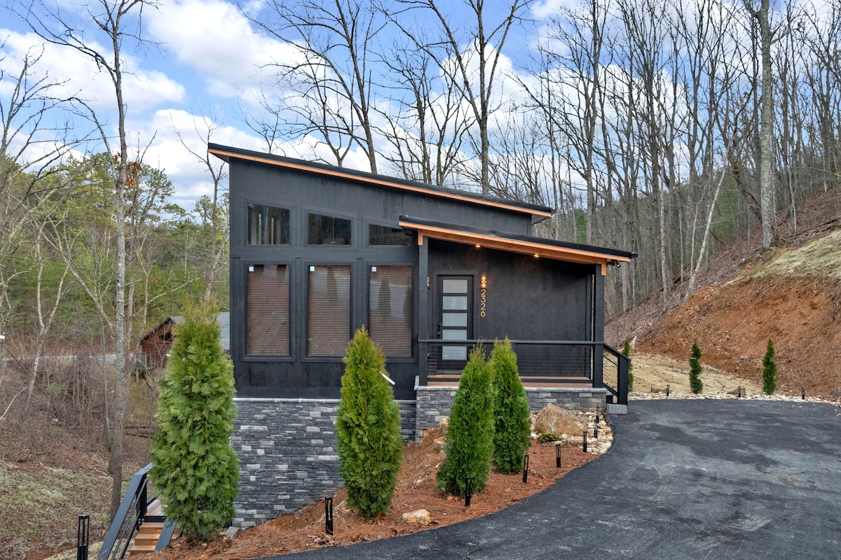 A modern cabin is presented within a serene wooded setting. The exterior features dark siding, large windows, and a welcoming porch. Lush green shrubs are positioned along the pathway, leading to the entrance. The surrounding landscape shows bare trees and soft earth tones of the hillside.