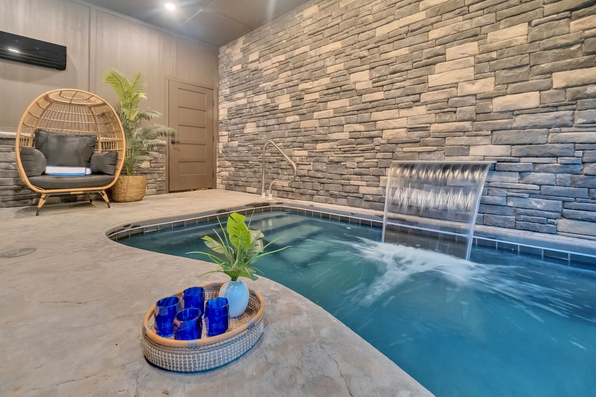 An indoor heated pool features soothing water flowing over a decorative stone wall. A wicker chair is positioned nearby, offering a cozy spot to relax. A tray with decorative glasses and a small plant adds a touch of elegance to the space.