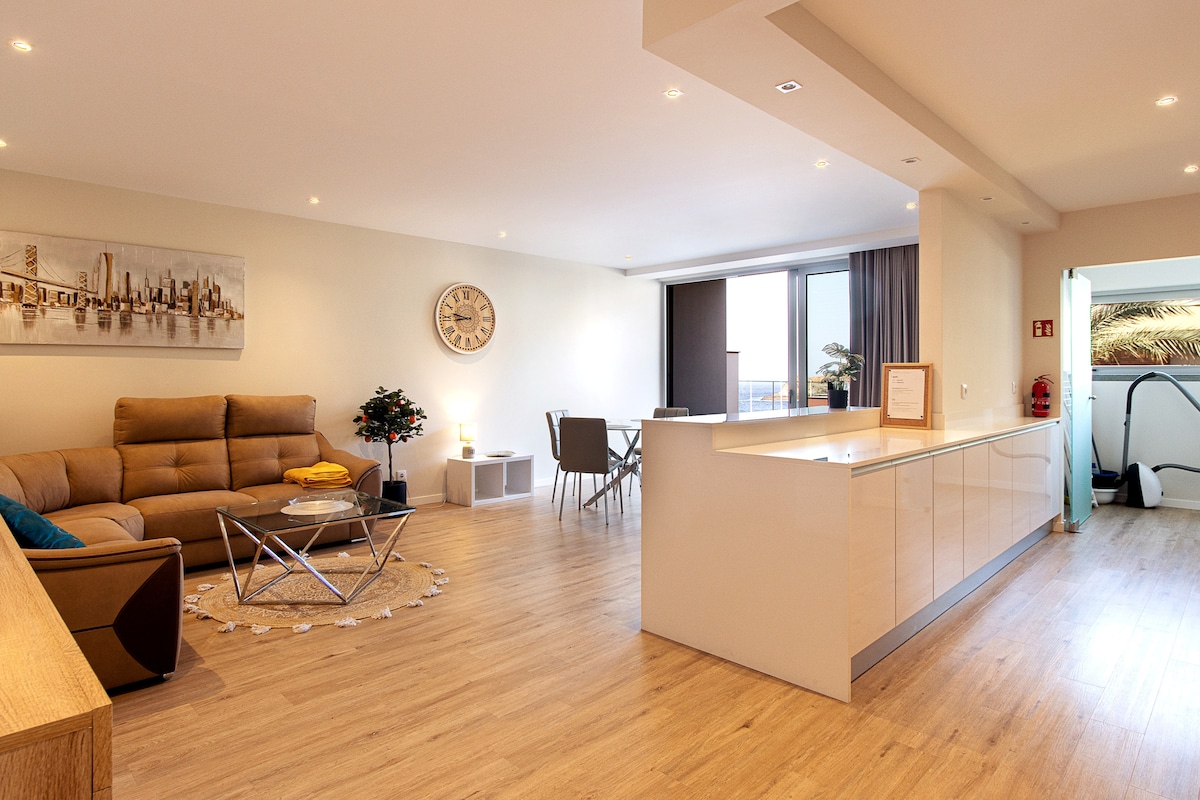 A spacious living and dining area features modern furnishings, including a comfortable sofa and a glass coffee table. Large windows provide ample natural light and showcase views of the outdoors. The open kitchen, equipped with contemporary cabinets and appliances, is visible in the background.
