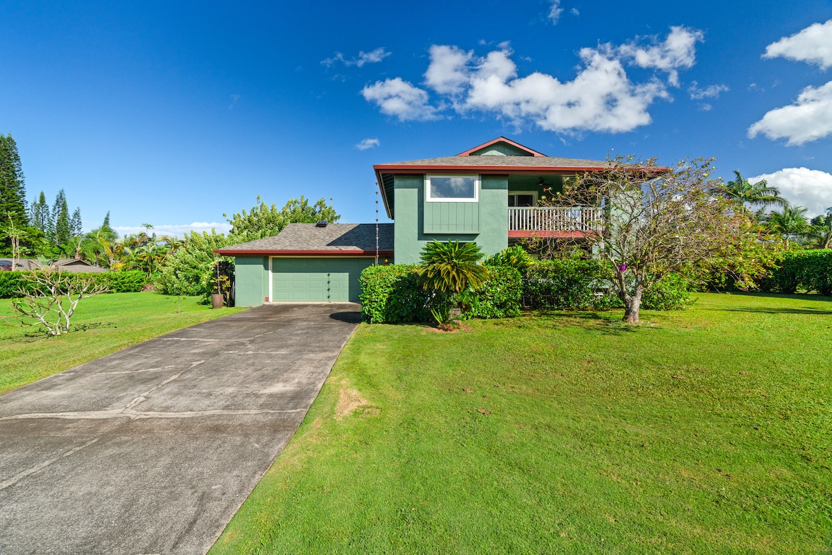A charming two-story home is shown set against a clear blue sky, surrounded by lush greenery. The driveway leads to a spacious garage, with tropical trees and plants framing the property, inviting a sense of tranquility and connection to nature.