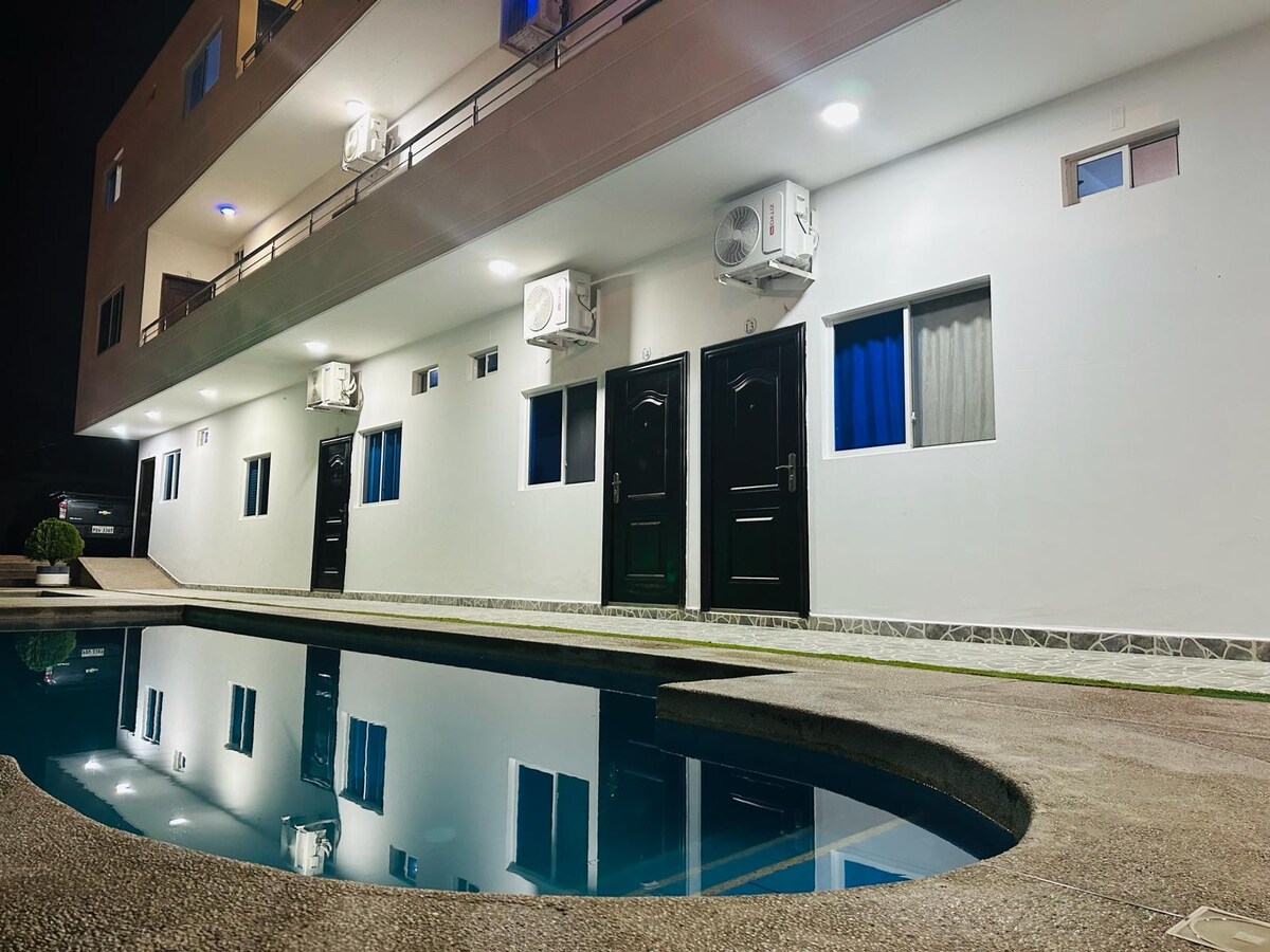 A modern building features multiple entrance doors with large windows above reflective pool waters. Air conditioning units are mounted on the exterior walls, while soft lighting illuminates the area, creating a calming evening ambiance.