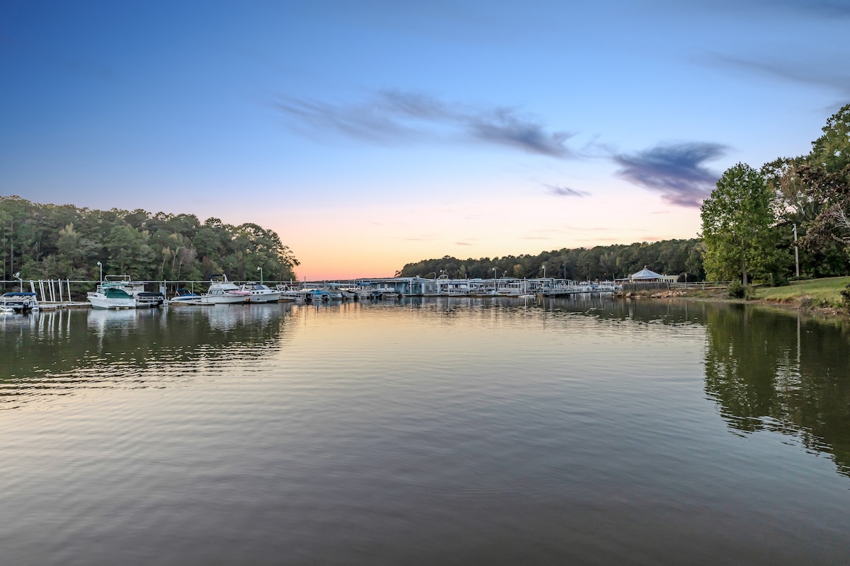 A calm lake scene is captured at dusk, reflecting hues of blue and soft pastel colors. Boats are moored at a dock, surrounded by lush green trees along the shore. The serene water creates a tranquil atmosphere, inviting moments of relaxation.