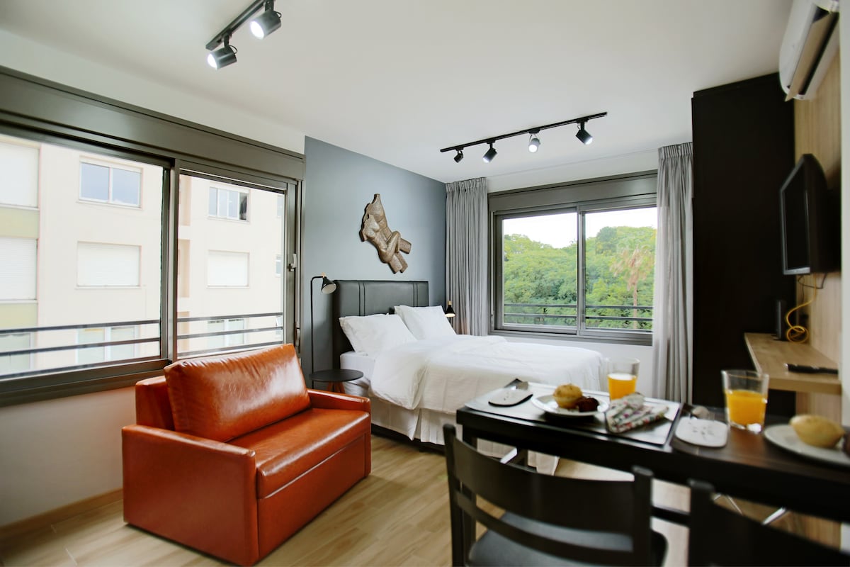 The room features a spacious bed accompanied by bedside lamps and large windows that offer a view of greenery outside. A cozy orange armchair sits beside a dining table with breakfast items, including juice and pastries. Natural light brightens the modern decor.
