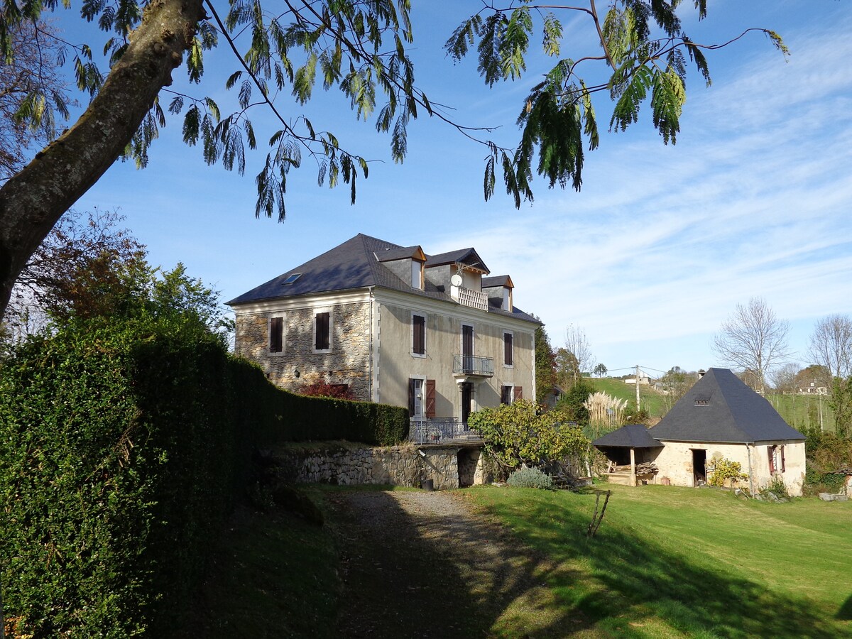 An elegant stone house is surrounded by lush greenery and open lawns. The structure features large windows and a symmetrical roofline. A garden area and an outbuilding are visible in the foreground, with a clear blue sky overhead.