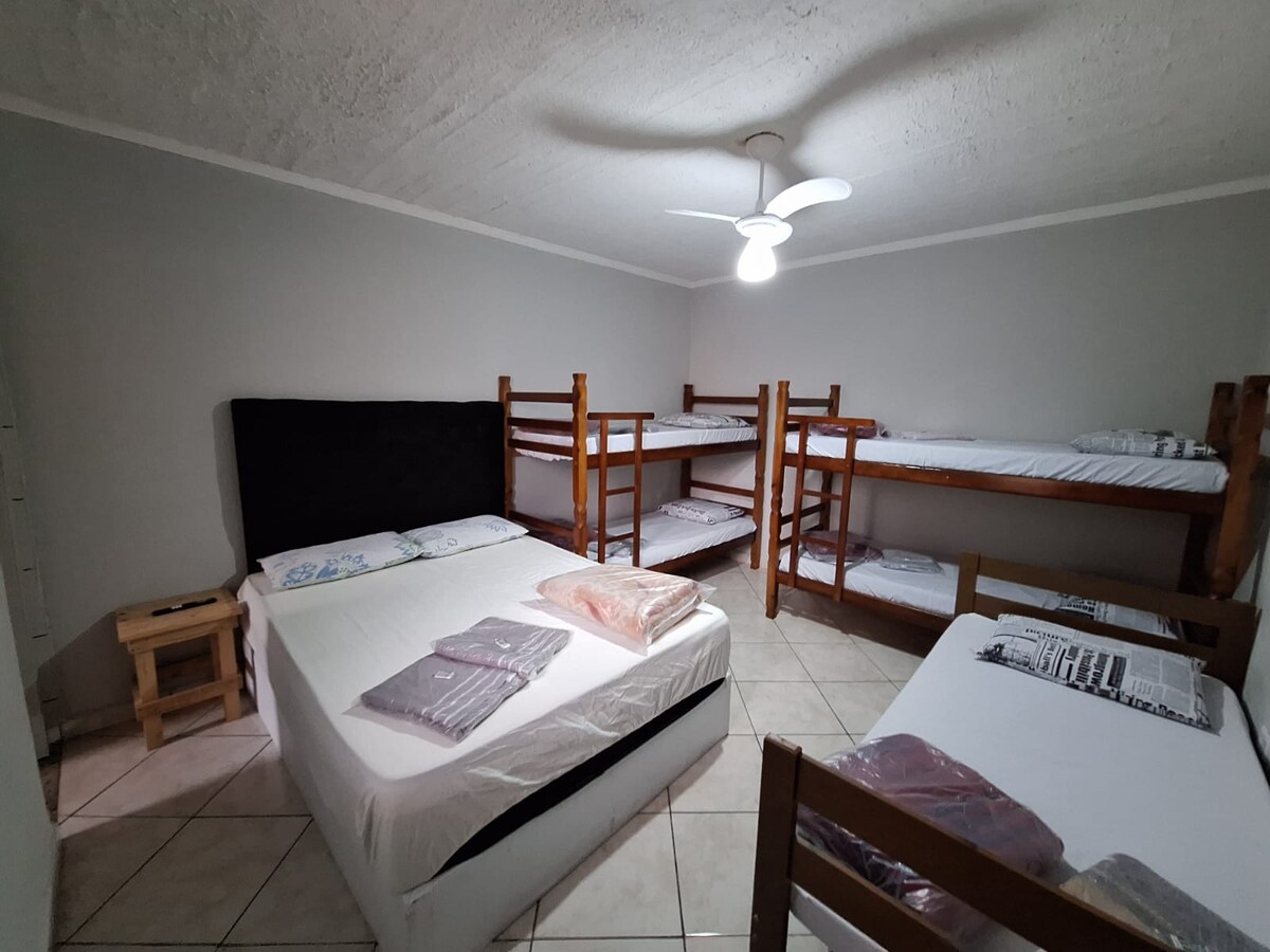 A private suite is shown featuring a spacious double bed and two single beds alongside a set of bunk beds. The room is equipped with a ceiling fan, while light tiles and neutral walls create a bright and open atmosphere.