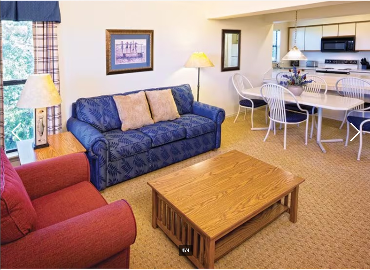 A living area features a blue sofa with decorative cushions, complemented by a red armchair. A wooden coffee table sits in front, surrounded by natural light from large windows. The dining space includes a round table with white chairs and a view of the kitchen area.
