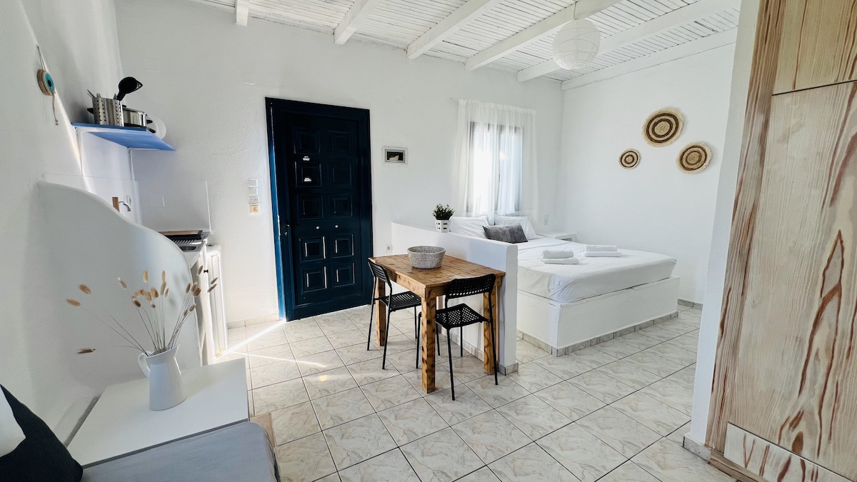 An inviting interior features a kitchenette, a wooden dining table with chairs, and a cozy sleeping area with a spacious bed. Natural light fills the room through a window, while decorative wall accents offer a touch of charm. The ceiling showcases exposed wooden beams.