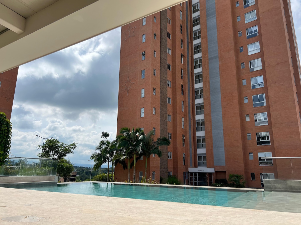 An outdoor infinity pool is framed by a marble deck, surrounded by lush greenery. The towering brick building stands in the background against a partly cloudy sky, providing a modern urban vibe. The tranquil water reflects the surrounding landscape, creating a serene atmosphere.