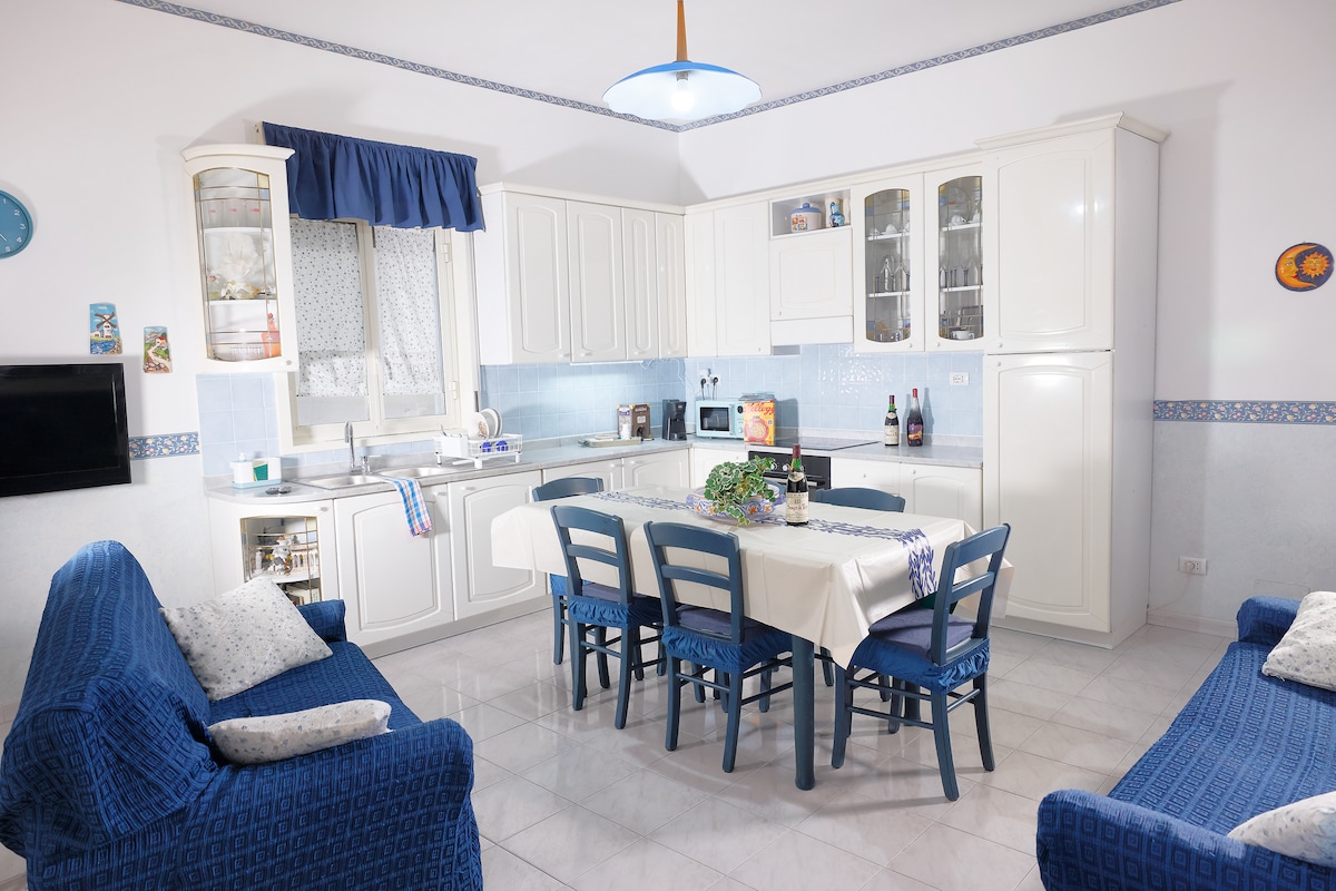A bright kitchen is presented with white cabinetry and blue accents. A dining table surrounded by chairs is centrally located, with a small plant as a centerpiece. Cozy sofas are positioned nearby, and a window allows natural light to illuminate the space.