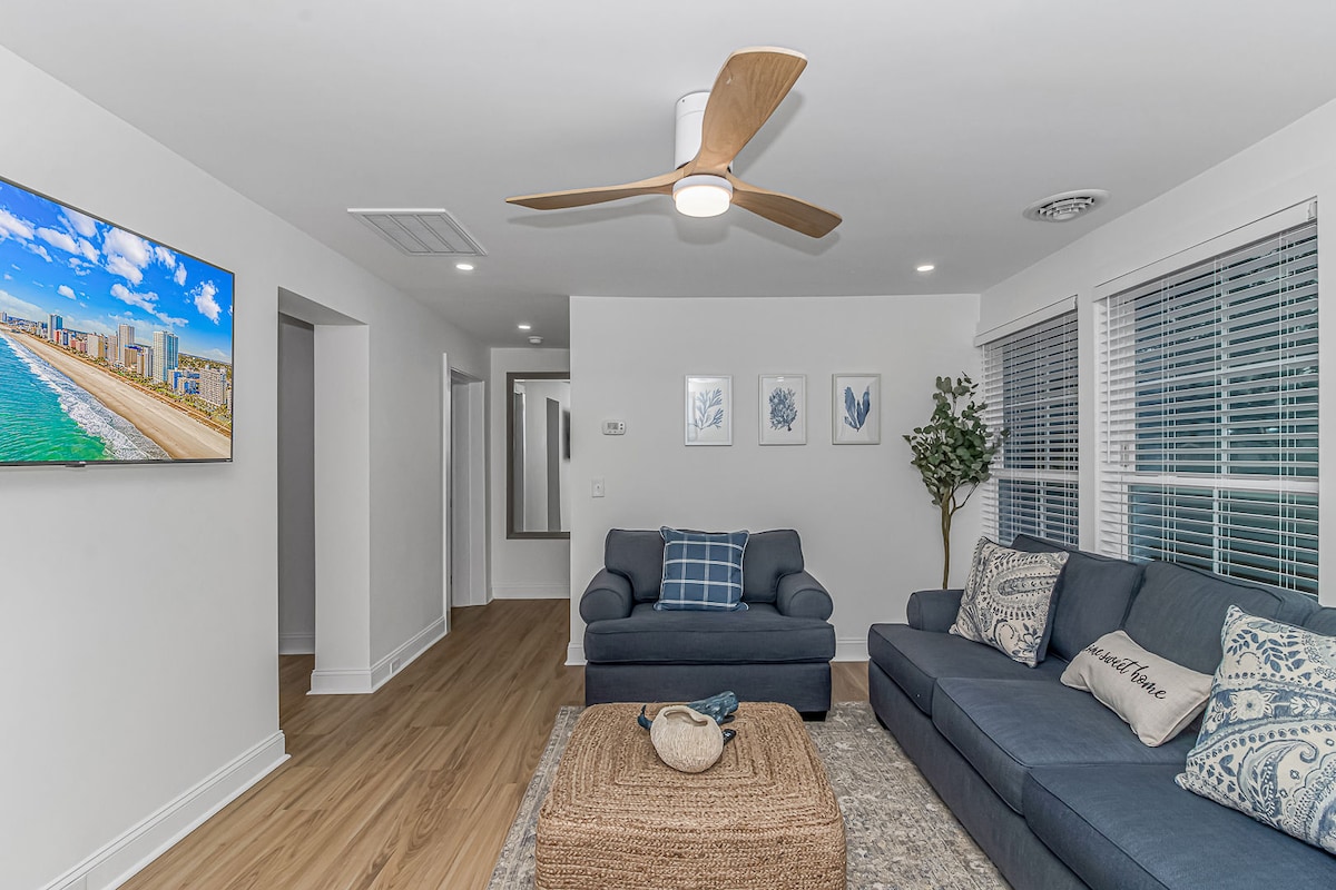 A modern living room features a comfortable blue sofa and armchair, arranged around a natural fiber area rug. A ceiling fan provides airflow, while a flat-screen television is mounted on the wall. Light filters through large windows adorned with blinds.