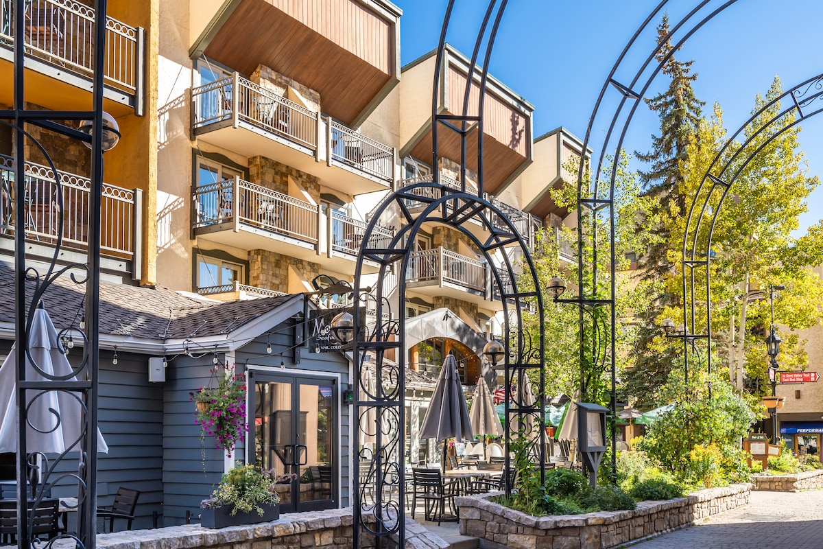 A charming courtyard is framed by an elegant archway structure, featuring shaded seating areas with umbrellas. The backdrop includes a series of multi-level buildings, each with balconies adorned with decorative railings. Lush greenery and vibrant flowers add to the serene environment.