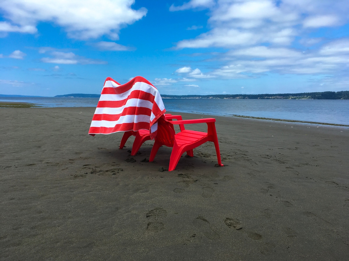 Two red lounge chairs are positioned on a sandy beach, with a striped blanket draped over one. The calm water extends to the horizon under a blue sky embellished with white clouds.