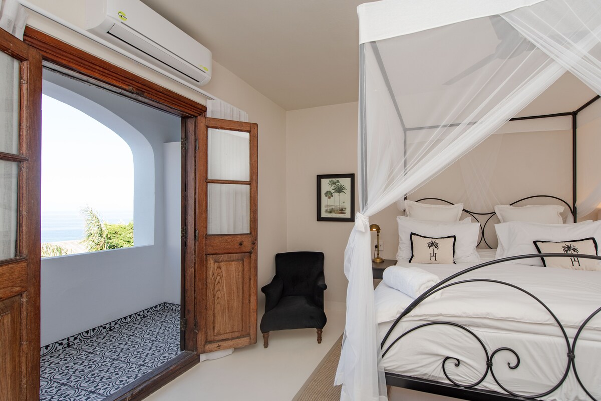 A spacious bedroom is presented with an elegant four-poster bed draped in a sheer mosquito net. A cozy chair is positioned near an open door, leading to a private balcony with ocean views. Bright sunlight illuminates the room, highlighting the intricate floor design.