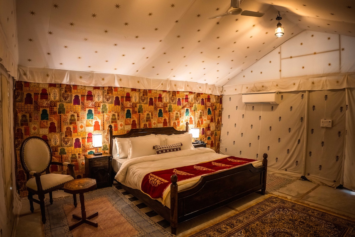 A spacious bedroom is adorned with intricate wall patterns featuring traditional motifs. The large bed is dressed in white linens, complemented by decorative blankets. Soft lighting is provided by bedside lamps, and a small table and chair are positioned nearby.