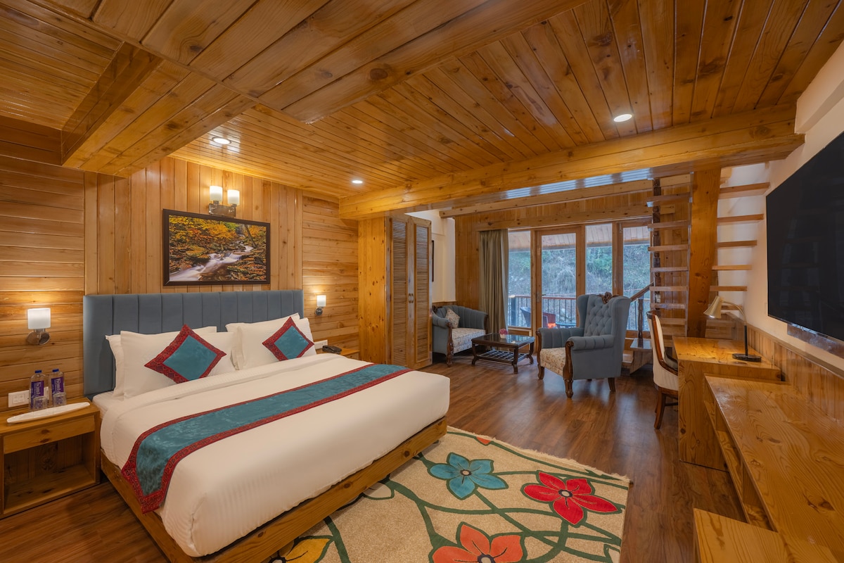 A wooden-paneled room features a spacious bed dressed in white linens with vibrant throw pillows. A colorful rug with floral patterns is situated on the floor. Cozy seating is provided by a pair of armchairs, and large windows offer views of the surrounding landscape.