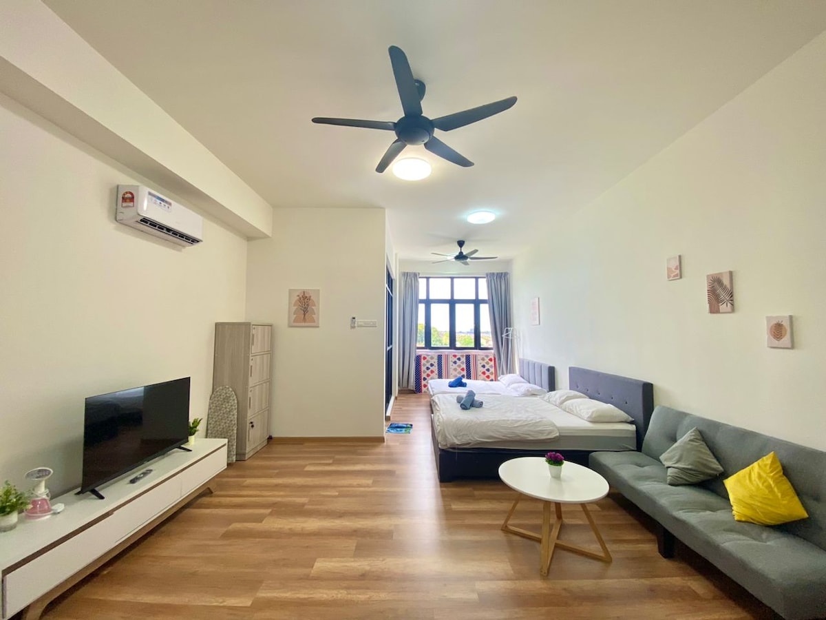 A spacious studio layout is presented with a large window allowing natural light to brighten the room. A comfortable bed is positioned beside a modern sofa. A small coffee table sits in the center, with a television mounted on a light-colored media console across the room.