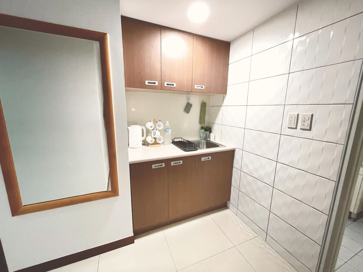 A compact kitchen area features modern cabinetry and a reflective mirror. Counter space includes a stovetop, kettle, and water bottles, complemented by light-colored tiles for a clean appearance. Bright overhead lighting enhances visibility, creating a functional cooking environment.
