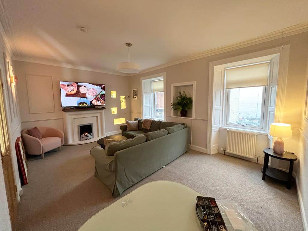 A spacious lounge area is furnished with a soft green sectional sofa, accompanied by a small armchair. Large windows allow natural light to fill the room, while a fireplace adds a cozy touch. A wall-mounted television is positioned for easy viewing.
