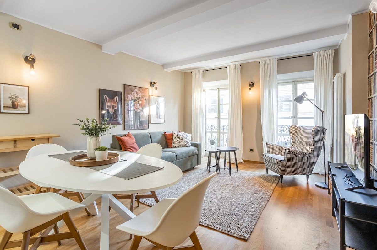 A bright living area is shown with a comfortable sofa, accent pillows, and a light-colored armchair. A round dining table with white chairs is positioned nearby. Large windows are covered with soft curtains, allowing natural light to fill the space. Wall art adds a touch of character.