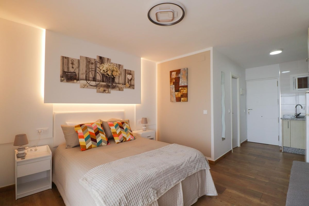 A well-lit sleeping area features a spacious bed with a light-colored coverlet. Colorful accent pillows add a pop of color against the neutral wall. The room includes two bedside tables with lamps and is connected to a kitchenette area visible in the background.