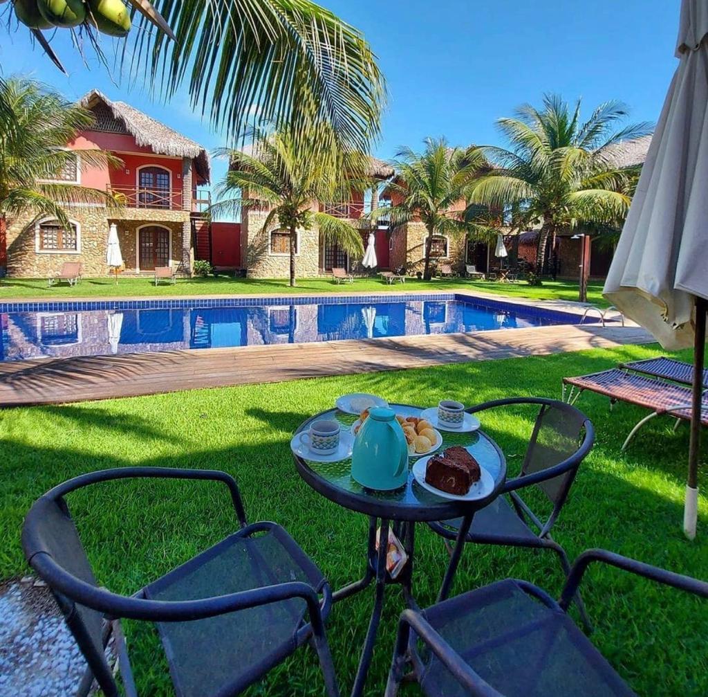 A pool area is presented, featuring a vibrant blue swimming pool surrounded by lush green grass. Two chairs and a small table are positioned nearby, set with a teapot and pastries. Palm trees provide a tropical backdrop, enhancing the outdoor leisure space.