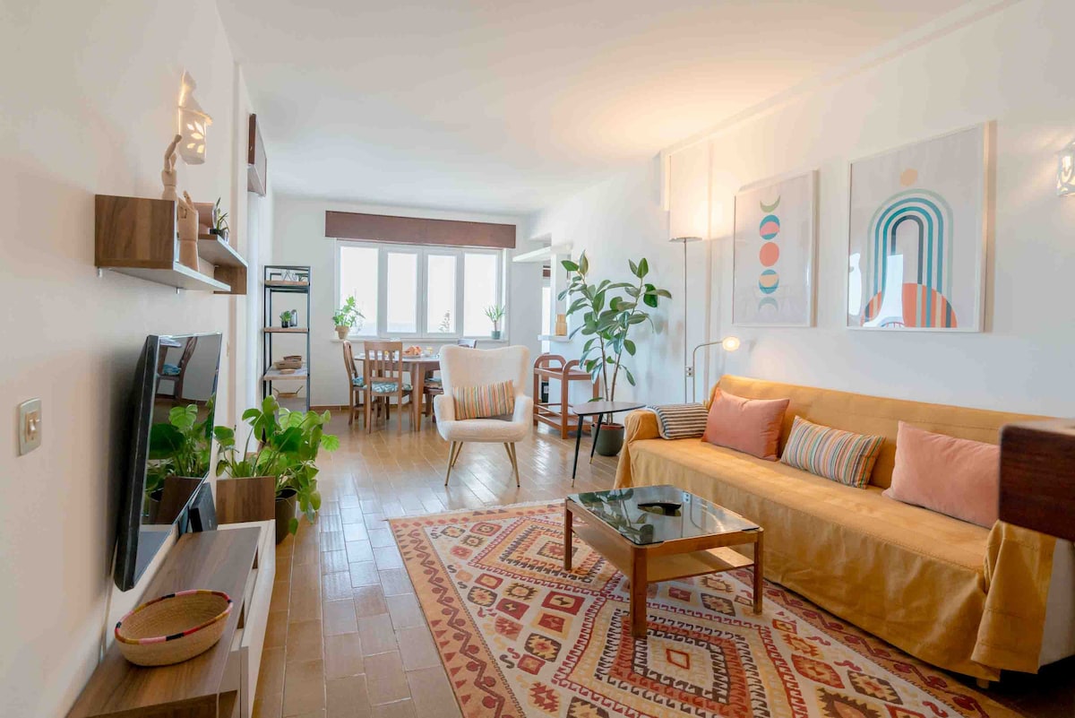 A bright living area features a spacious sofa adorned with cushions, a glass coffee table, and a television mounted on the wall. Artistic wall hangings add interest. Potted plants enhance the natural feel, while a dining area is visible in the background, illuminated by natural light.