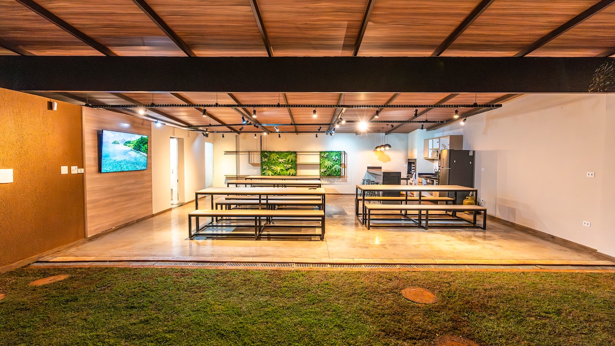A spacious indoor area features multiple long tables arranged for gatherings, complemented by modern lighting. Large windows offer views of the surrounding green grass. A kitchen area is visible in the background, along with colorful wall art that adds a fresh touch to the space.