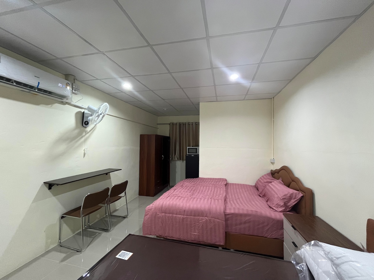 A spacious room is furnished with two beds covered in light pink bedding. A small dining table with two chairs is positioned near the entrance. Additional furniture includes a dresser and a wardrobe, while natural light enters through a partially covered window.