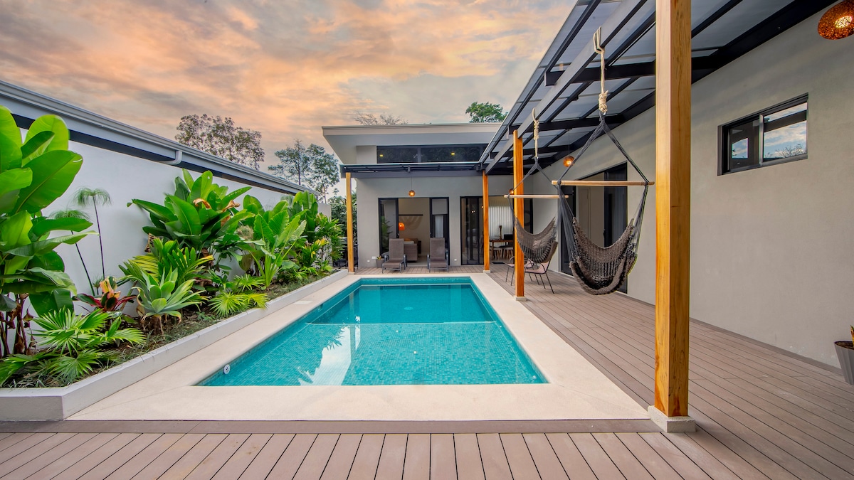 An inviting outdoor area is highlighted by a crystal-clear pool surrounded by wooden decking and lush greenery. Two hammocks are suspended nearby, and comfortable seating can be seen beneath a covered patio, providing a serene space for relaxation amidst a tropical atmosphere.