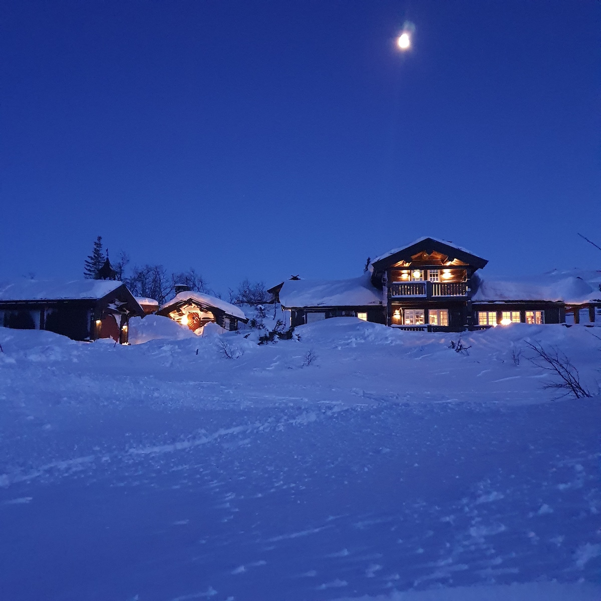 A mountain lodge is depicted under a clear night sky, with a bright moon illuminating the surroundings. Snow blankets the terrain, and warmly lit windows suggest a cozy interior. The structure features multiple sections, with a gently sloping roof and wooden accents visible.
