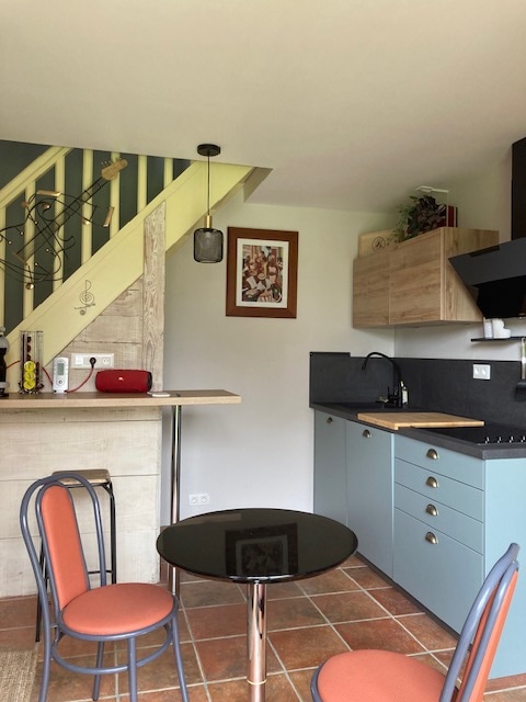 The kitchen area features modern cabinetry in soft hues, complemented by a black countertop. A small round table with two chairs is positioned nearby, while a staircase leads to the upper level. Wall art offers a touch of decor, enhancing the practical space.