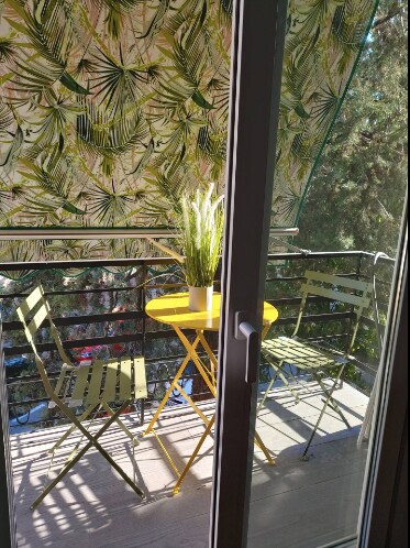 A small balcony is depicted, featuring a round yellow table surrounded by two green folding chairs. A potted plant sits on the table, and a vibrant patterned awning provides shade. The scene showcases a tranquil outdoor space, accessible from the interior.