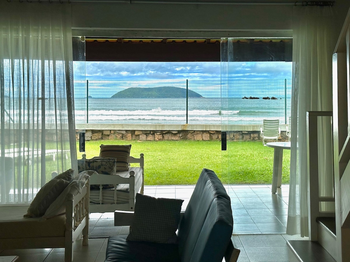 A spacious living area offers a clear view of the ocean through large glass doors, framed by sheer white curtains. The lush green lawn and sandy beach are visible beyond a protective fence, creating a seamless connection between indoor and outdoor spaces.