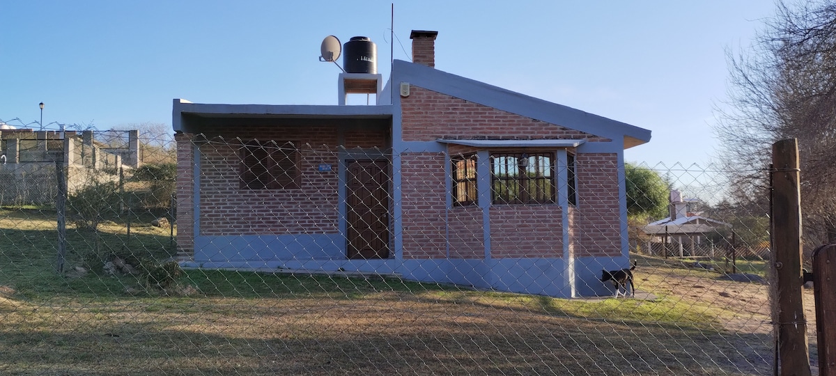 A single-story house is presented with a red brick exterior and light blue accents. Several windows allow natural light inside, and a water tank is mounted on the roof. A grassy area is visible in front, with a dog standing beside the house.