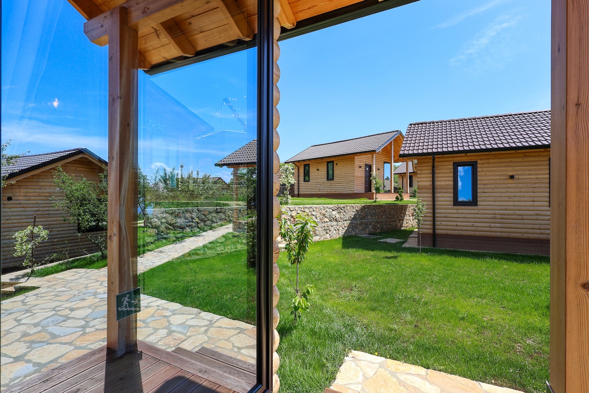 A clear view from a glass doorway reveals a well-maintained outdoor space featuring stone paths and lush greenery. Quiet wooden cabins are positioned on a gentle slope, surrounded by manicured lawns, under a bright blue sky.
