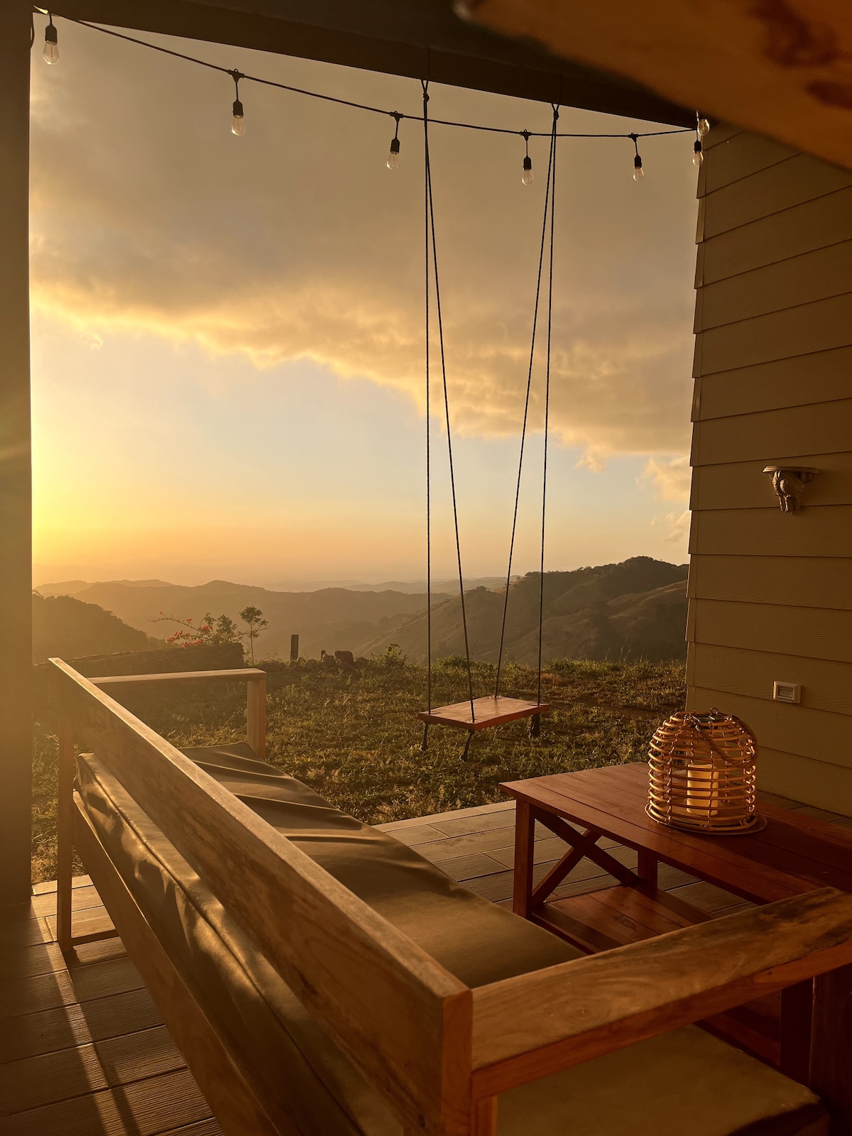 A private terrace features a wooden bench set against a backdrop of mountains and a sunset sky. A swing hangs from the overhang, while a decorative lantern rests on a small table, casting a warm glow. Natural beauty is highlighted in the serene setting.