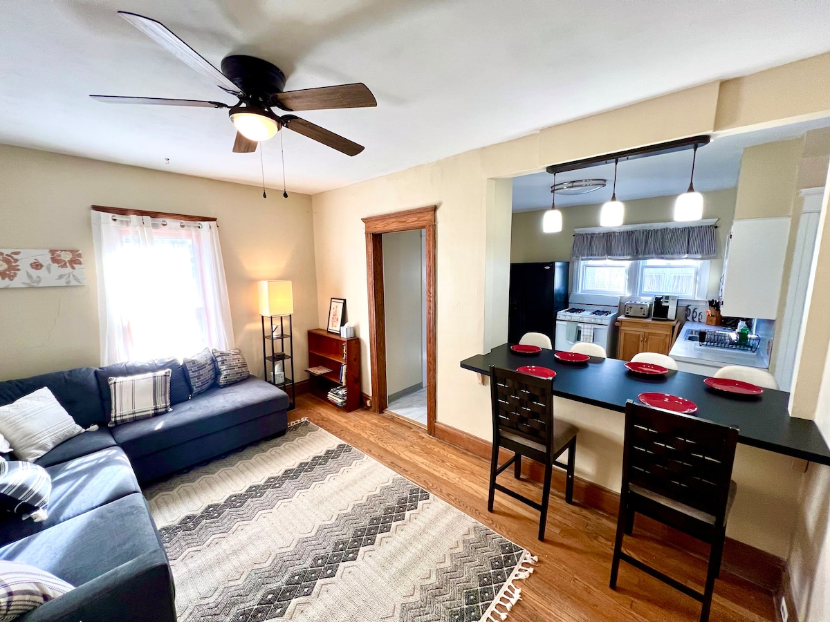The living area showcases a spacious blue sofa accompanied by a neutral area rug. A dining bar with two chairs is positioned near the kitchen entrance. Natural light filters through a window adorned with light curtains, enhancing the cozy ambiance.
