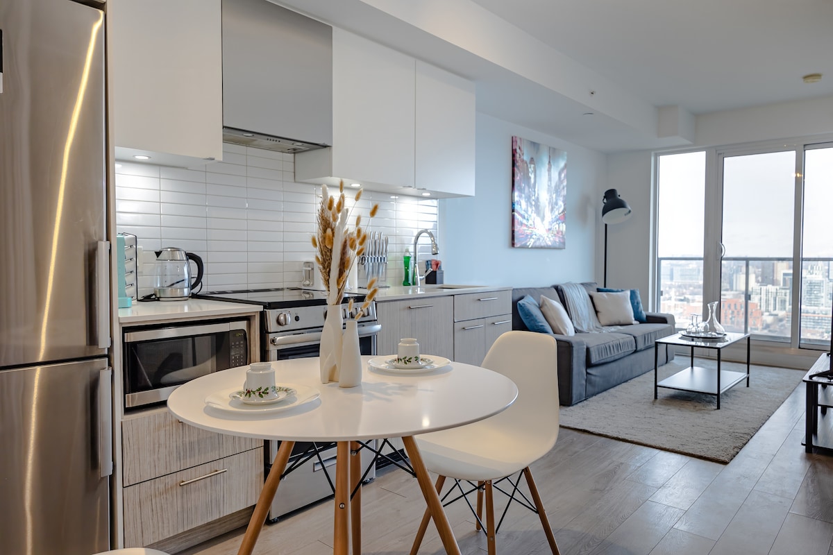 A modern kitchen area features sleek cabinetry and stainless-steel appliances, including a stove and refrigerator. A round dining table with white chairs is set for two. The living space includes a comfortable sectional sofa, a coffee table, and large windows offering city views.