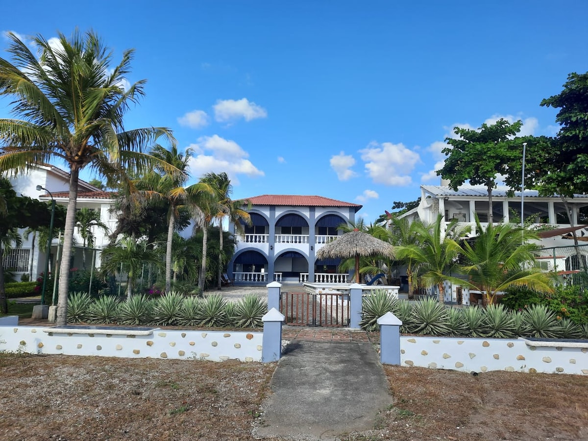 The residence features a charming two-story design with a red-tiled roof and arched balconies. Lush palm trees and well-kept landscaping surround the property, while a pathway leads to the entrance. A thatched-roof gazebo adds to the inviting outdoor atmosphere.