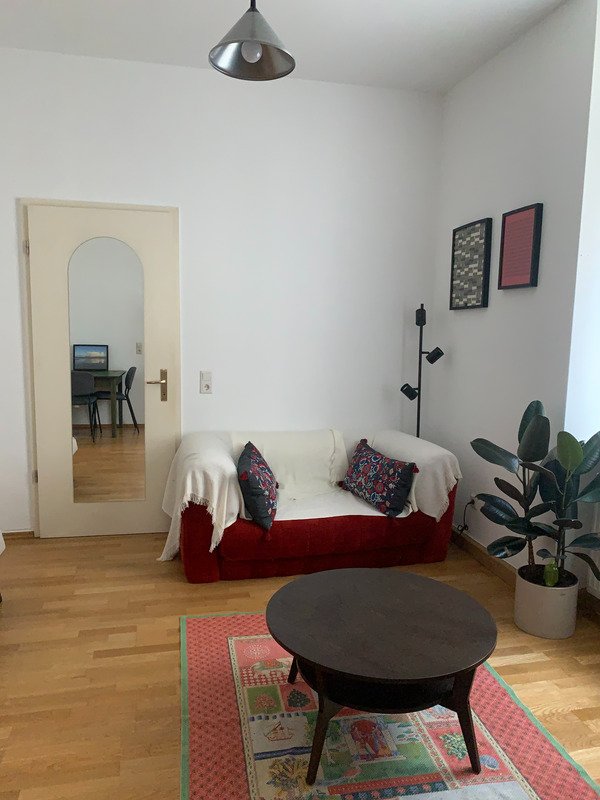 A cozy seating area features a red couch adorned with patterned cushions, complemented by a decorative rug. A circular coffee table occupies the center, while a plant adds a touch of greenery. A partially visible door opens into another space, suggesting connectivity.