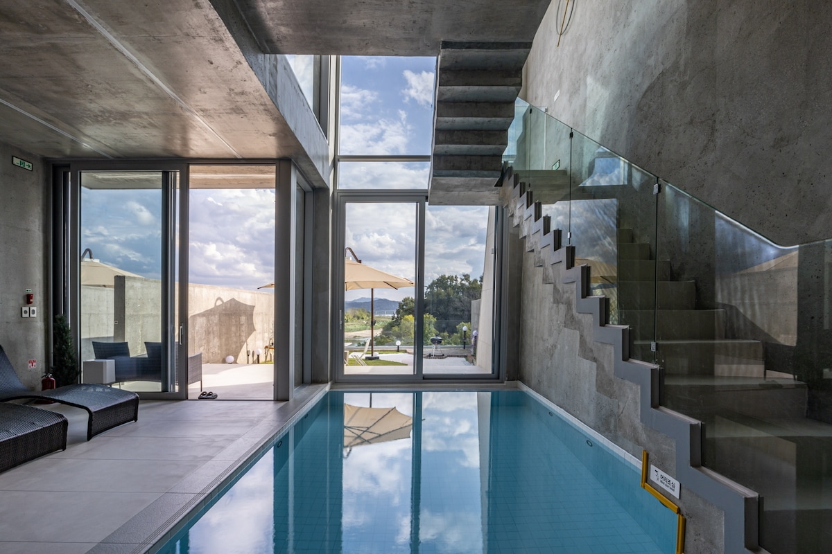 An indoor infinity pool is framed by large glass windows, allowing natural light to fill the space. A staircase adorned with sleek glass railings leads to the upper level, creating an open and airy atmosphere. Views of the outdoor area can be seen through the glass doors.