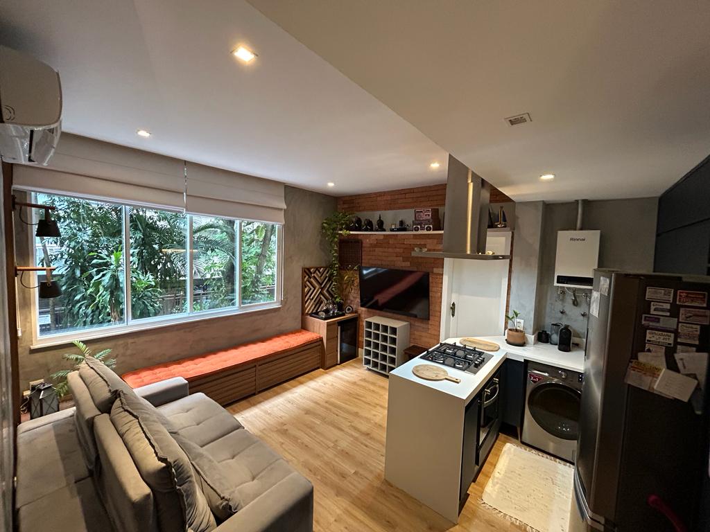 An open-plan living space includes a comfortable gray sofa and a wall-mounted television. Light fills the room through large windows overlooking greenery. A modern kitchen with a gas stove and a refrigerator is visible, complemented by wooden cabinets and a small dining area.