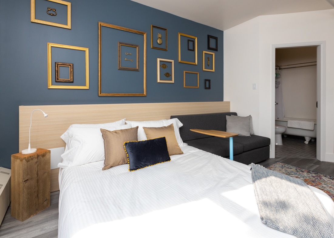 A studio space features a plush bed with decorative pillows, complemented by a sleek couch with a side table. A wall adorned with an arrangement of empty frames adds character, while a doorway leads to a bathroom with a claw-foot tub visible.