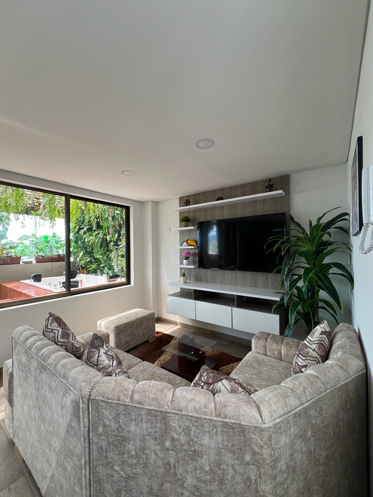 A living area is presented, featuring a comfortable, light-colored sofa accompanied by two armchairs. A coffee table occupies the center space, and a wall-mounted television is visible. Large windows allow natural light, while greenery is seen outside, providing a view of the surrounding area.