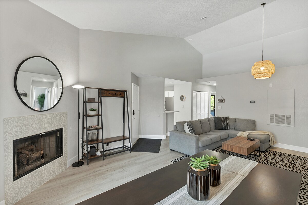 A stylish living area features a modern sectional sofa paired with a wooden coffee table. Natural light floods the space through large windows, while a round mirror and decorative shelving unit add an inviting touch. A minimalist fireplace enhances the ambiance of the room.