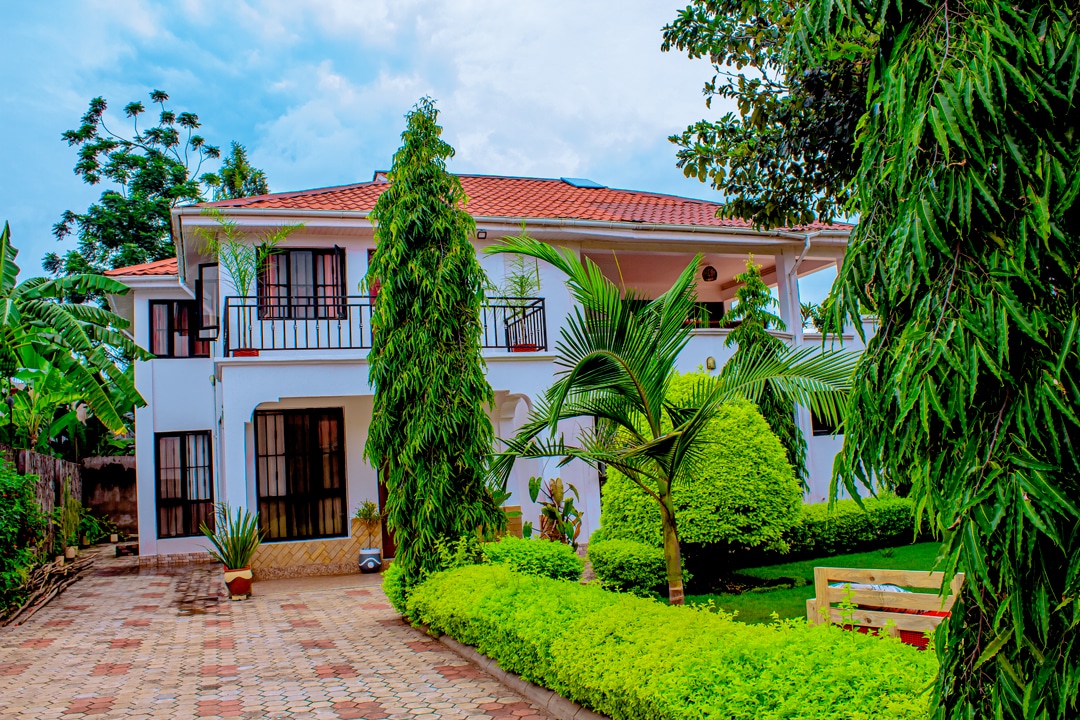 The exterior of a two-story house is framed by lush greenery, featuring manicured lawns and tall hedges. Paved pathways lead to the entrance, while an inviting porch and balcony offer views of the surrounding garden, creating a serene and welcoming atmosphere.