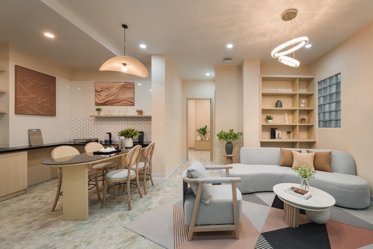 A spacious living area features a blend of modern furniture, with a light-colored sofa and a round coffee table at the center. The kitchen area, visible in the background, includes sleek cabinetry and a dining table surrounded by stylish chairs, with decorative elements displayed on shelves.